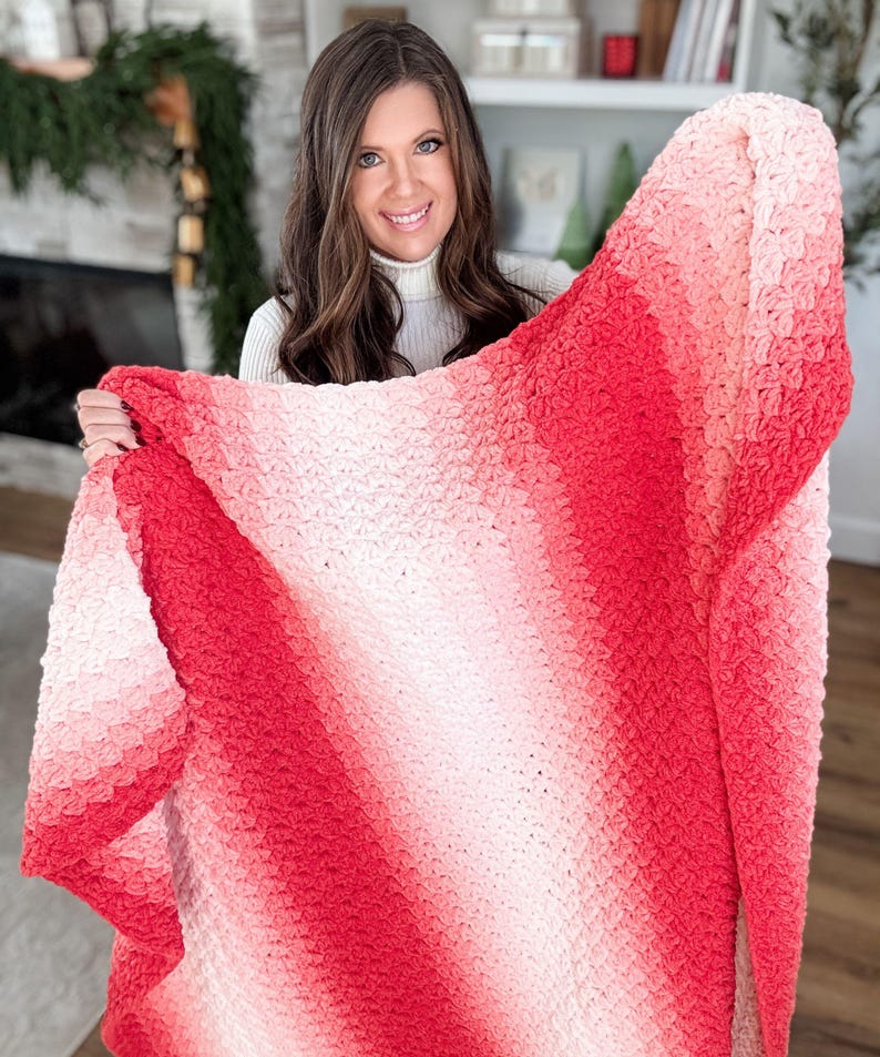 May include: A handmade, crochet blanket with a gradient of colors from light pink to deep red. The blanket has a textured stitch pattern and is displayed against a neutral background. The blanket is a cozy home decor item.