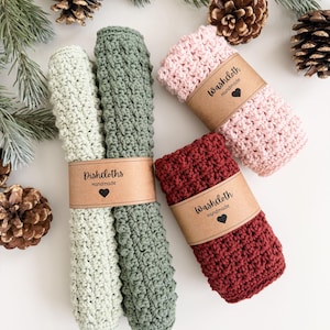 May include: Four handmade crochet washcloths in shades of green, pink, and burgundy, each wrapped with a brown paper band. The green washcloths are labeled "Dishcloths Handmade" and the others are labeled "Washcloth Handmade". Pinecones and evergreen branches are in the background.