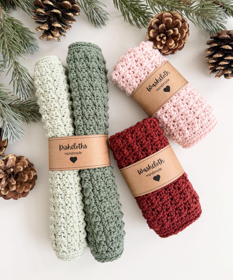 May include: Four handmade crochet washcloths in shades of green, pink, and burgundy, each wrapped with a brown paper band. The green washcloths are labeled "Dishcloths Handmade" and the others are labeled "Washcloth Handmade". Pinecones and evergreen branches are in the background.