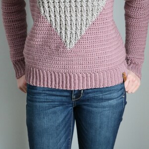 Puede incluir: Primer plano de un su&eacute;ter de crochet rosa con un dise&ntilde;o de tri&aacute;ngulo de crochet gris. El su&eacute;ter se lleva sobre unos vaqueros azules.
