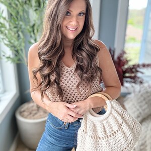 May include: A woman is holding a white crocheted tote bag with a wooden handle. The bag has a large open top and a woven design. The woman is wearing a brown crochet top and blue jeans.