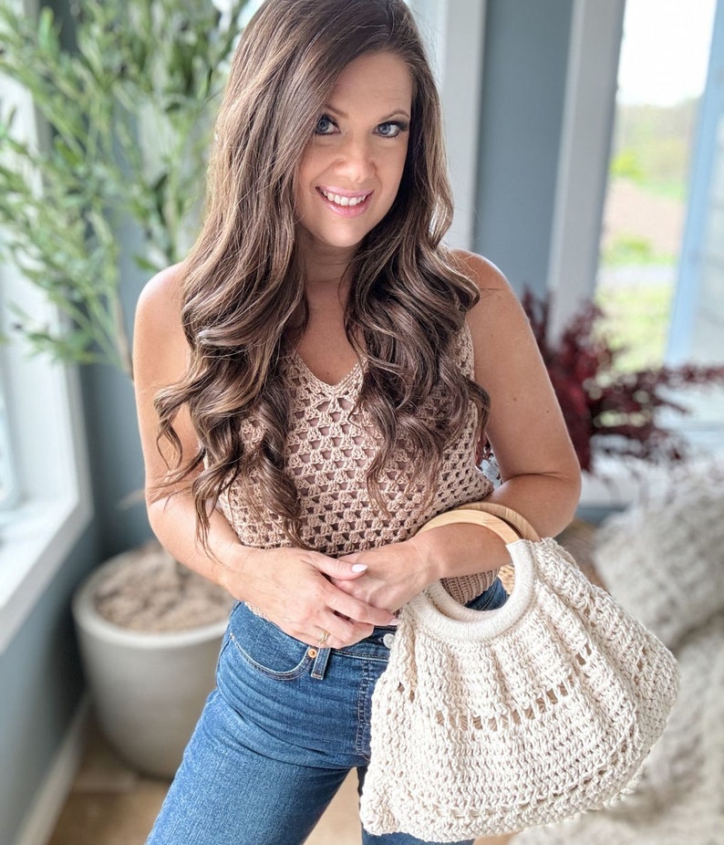 May include: A woman is holding a white crocheted tote bag with a wooden handle. The bag has a large open top and a woven design. The woman is wearing a brown crochet top and blue jeans.