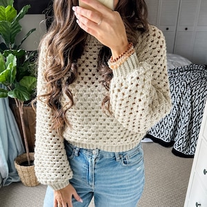 Pu&ograve; includere: Un maglione all'uncinetto color crema con un design a trama larga e aperta. Il maglione ha maniche lunghe e voluminose e un polsino a coste. La modella indossa jeans blu. Il maglione &egrave; un capo elegante e confortevole.