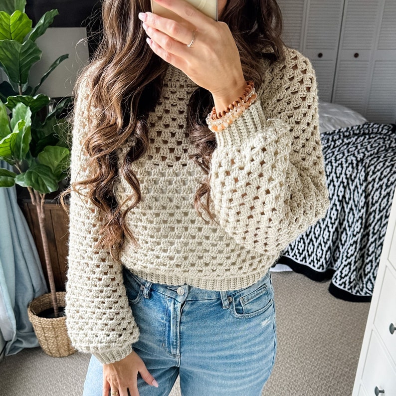 Pu&ograve; includere: Un maglione all'uncinetto color crema con un design a trama larga e aperta. Il maglione ha maniche lunghe e voluminose e un polsino a coste. La modella indossa jeans blu. Il maglione &egrave; un capo elegante e confortevole.