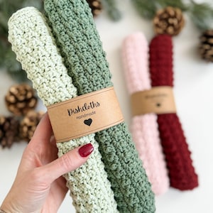 May include: Handmade crochet dishcloths in light green, green, pink, and burgundy. Each pair is wrapped with a brown paper band that reads "Dishcloths Handmade" with a heart symbol. The dishcloths are made of a textured, absorbent material.