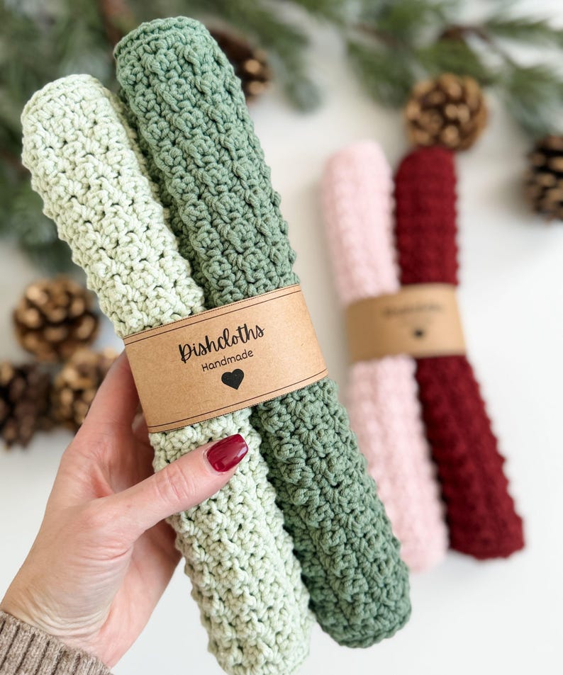 May include: Handmade crochet dishcloths in light green, green, pink, and burgundy. Each pair is wrapped with a brown paper band that reads "Dishcloths Handmade" with a heart symbol. The dishcloths are made of a textured, absorbent material.