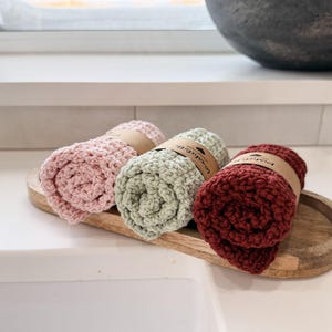 May include: Three rolled washcloths in pink, sage green, and burgundy, each secured with a brown paper band. The washcloths are displayed on a wooden tray, showcasing their textured, handmade appearance. The background includes a white countertop and a window.