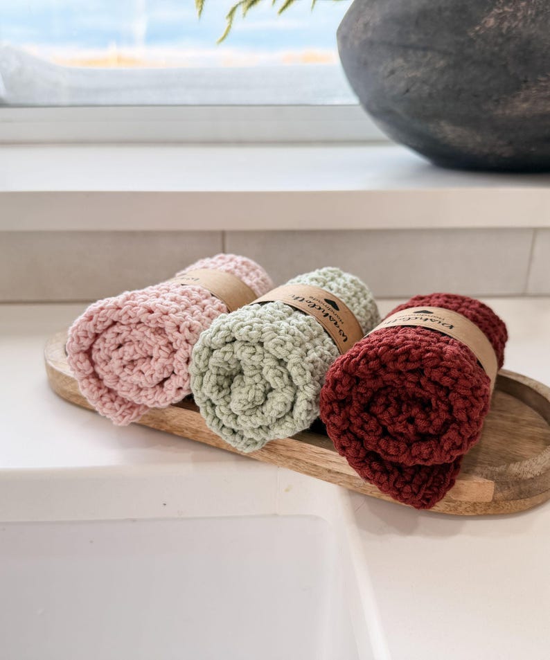 May include: Three rolled washcloths in pink, sage green, and burgundy, each secured with a brown paper band. The washcloths are displayed on a wooden tray, showcasing their textured, handmade appearance. The background includes a white countertop and a window.