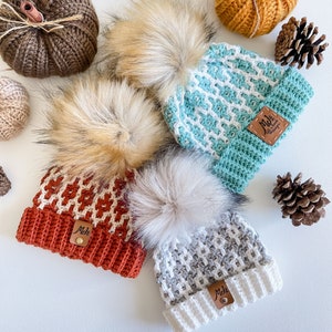 May include: Three crocheted hats with faux fur pom poms. The hats are in teal, orange, and gray with white accents. Each hat has a brown leather tag with the text "MJs Handmade" on it.