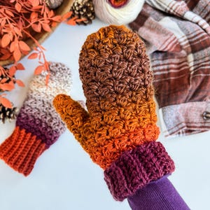 Peut inclure: Mitaines crochet&eacute;es &agrave; la main, aux couleurs automnales : orange, marron et violet. Les mitaines pr&eacute;sentent un point textur&eacute; et un poignet c&ocirc;tel&eacute;. L'une est port&eacute;e sur une manche violette. L'arri&egrave;re-plan comprend de la laine et du feuillage d'automne.