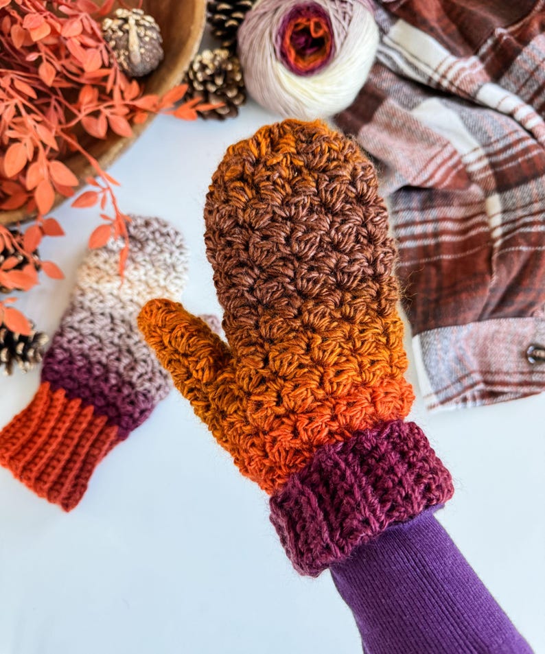 Peut inclure: Mitaines crochet&eacute;es &agrave; la main, aux couleurs automnales : orange, marron et violet. Les mitaines pr&eacute;sentent un point textur&eacute; et un poignet c&ocirc;tel&eacute;. L'une est port&eacute;e sur une manche violette. L'arri&egrave;re-plan comprend de la laine et du feuillage d'automne.
