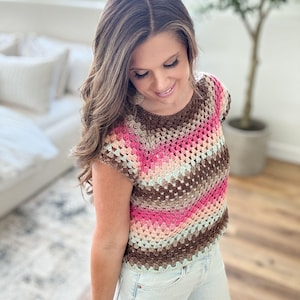 May include: A colorful crochet top in shades of brown, pink, and light blue. The short-sleeved top has a round neckline and a textured, open weave pattern. The top is paired with light-wash denim jeans.