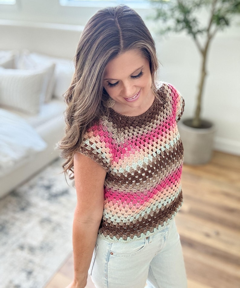 May include: A colorful crochet top in shades of brown, pink, and light blue. The short-sleeved top has a round neckline and a textured, open weave pattern. The top is paired with light-wash denim jeans.