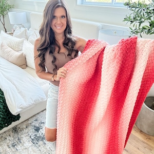 May include: A soft, textured blanket with a gradient of red and pink hues. The blanket is held up, showcasing its plush, woven design. The color transitions from light pink to a deep red, creating a visually appealing effect. This cozy blanket is perfect for home decor.