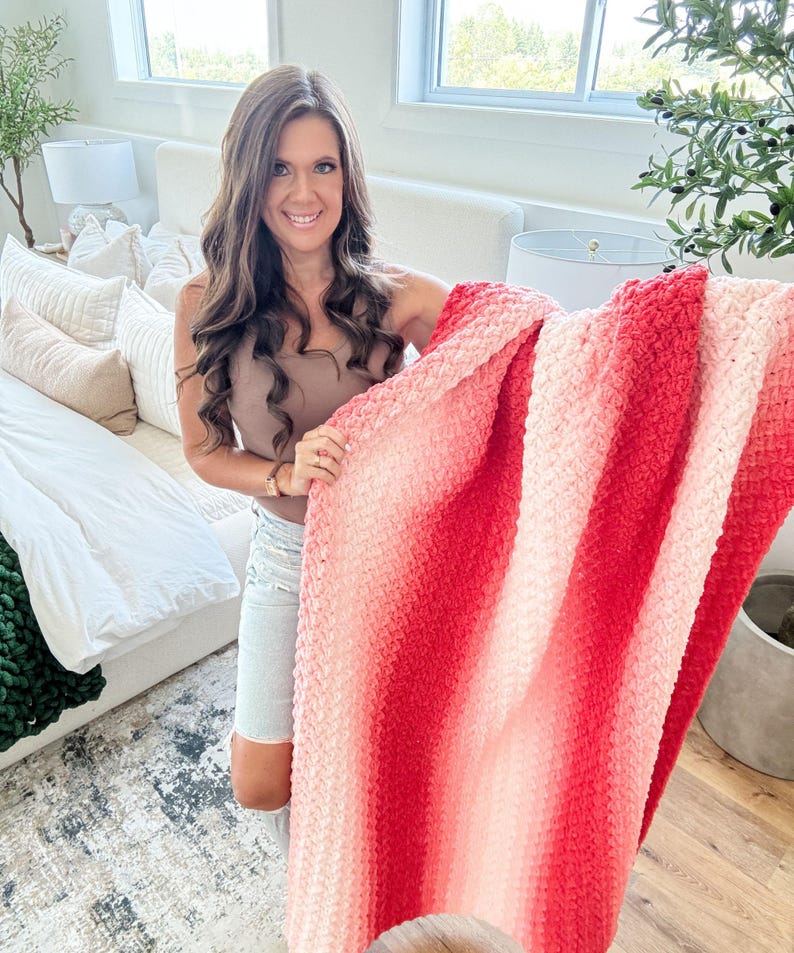 May include: A soft, textured blanket with a gradient of red and pink hues. The blanket is held up, showcasing its plush, woven design. The color transitions from light pink to a deep red, creating a visually appealing effect. This cozy blanket is perfect for home decor.