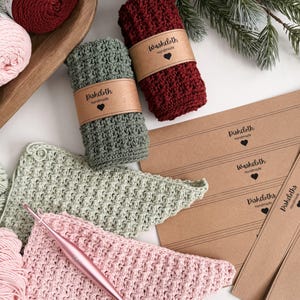 May include: Handmade crochet dishcloths and washcloths in shades of green, burgundy, and pink. The dishcloths are rolled and wrapped with a brown paper label that reads "Dishcloth Handmade" and "Washcloth Handmade". A crochet hook and yarn are also visible.