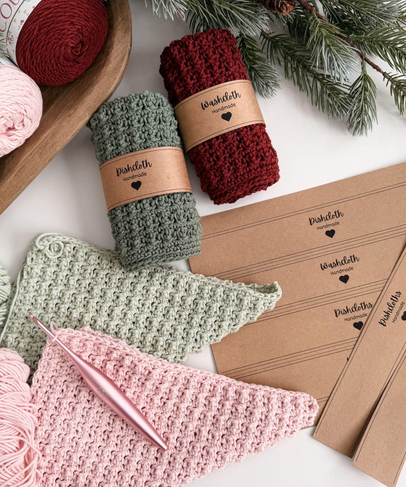 May include: Handmade crochet dishcloths and washcloths in shades of green, burgundy, and pink. The dishcloths are rolled and wrapped with a brown paper label that reads "Dishcloth Handmade" and "Washcloth Handmade". A crochet hook and yarn are also visible.