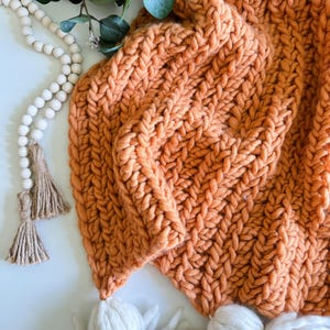 Puede incluir: Una manta de punto grueso de color naranja quemado con ribete de pompones blancos. La manta está doblada y drapeada sobre una superficie blanca con una guirnalda de cuentas de madera y borlas.