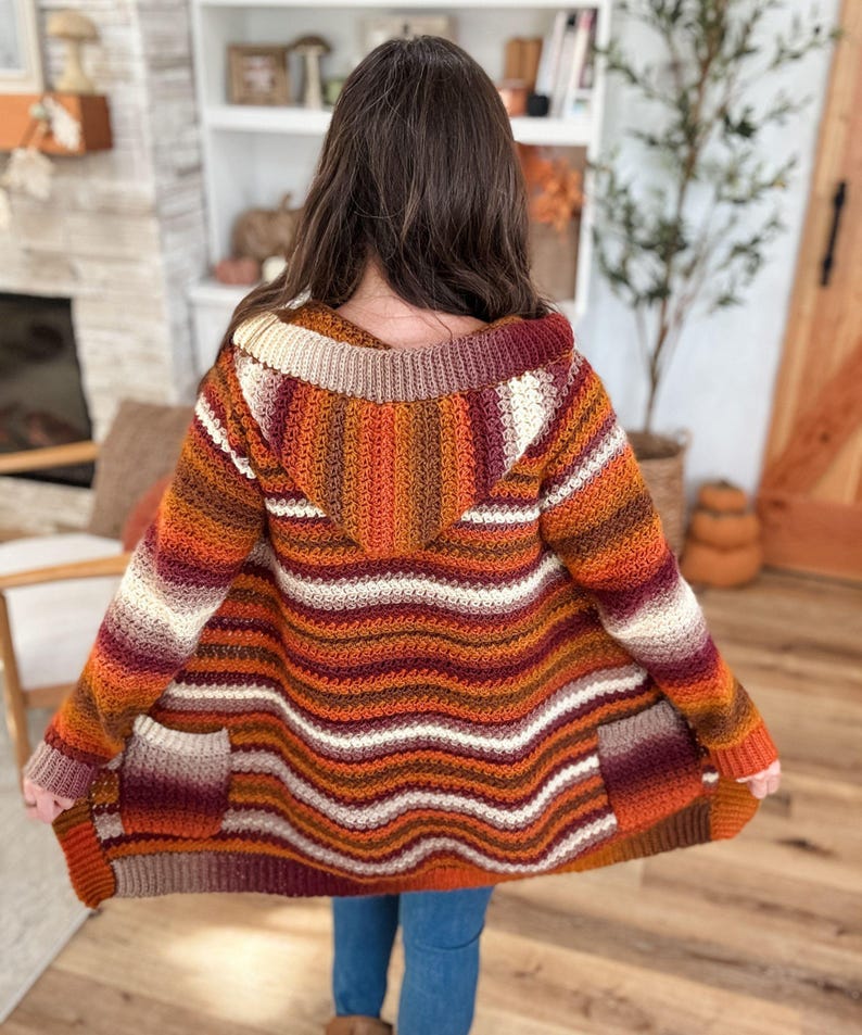 May include: A hooded, open-front cardigan in a horizontal striped pattern. The color palette includes shades of orange, burgundy, cream, and brown. The cardigan has two front pockets and is made of a textured, possibly crocheted, material.
