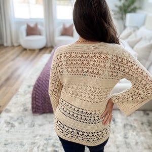 K&ouml;nnte beinhalten: Ein cremefarbener H&auml;kelpullover mit langen, ausgestellten &Auml;rmeln. Der Pullover hat ein offenes, spitzenartiges Design auf dem R&uuml;cken und im unteren Bereich, mit einem durchgehenden Band &uuml;ber dem Mittelteil. Das Kleidungsst&uuml;ck wird mit dunkelblauen Jeans getragen.