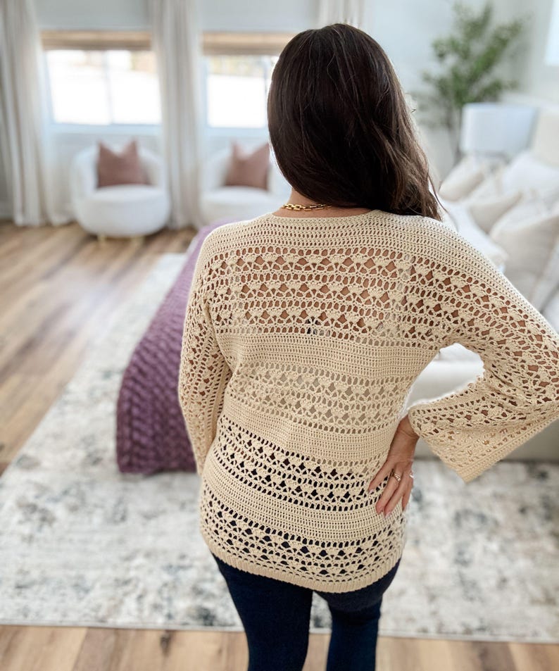 K&ouml;nnte beinhalten: Ein cremefarbener H&auml;kelpullover mit langen, ausgestellten &Auml;rmeln. Der Pullover hat ein offenes, spitzenartiges Design auf dem R&uuml;cken und im unteren Bereich, mit einem durchgehenden Band &uuml;ber dem Mittelteil. Das Kleidungsst&uuml;ck wird mit dunkelblauen Jeans getragen.