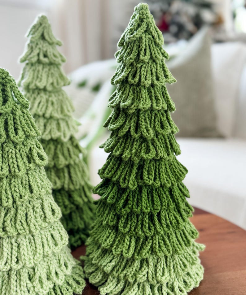 May include: Three handmade, knitted Christmas trees in varying shades of green. The trees are arranged on a wooden surface, with a soft, blurred background. The knitted texture creates a unique, textured appearance, perfect for holiday decor.