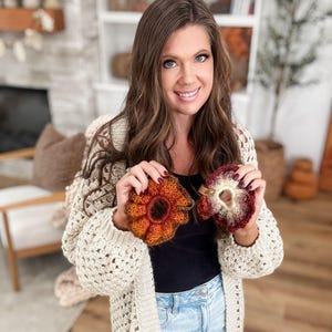 May include: Two handmade crochet hair scrunchies in shades of orange, red, and cream are held up. The scrunchies have a ruffled, flower-like design. The person holding the scrunchies is wearing a cream-colored, open-knit cardigan.