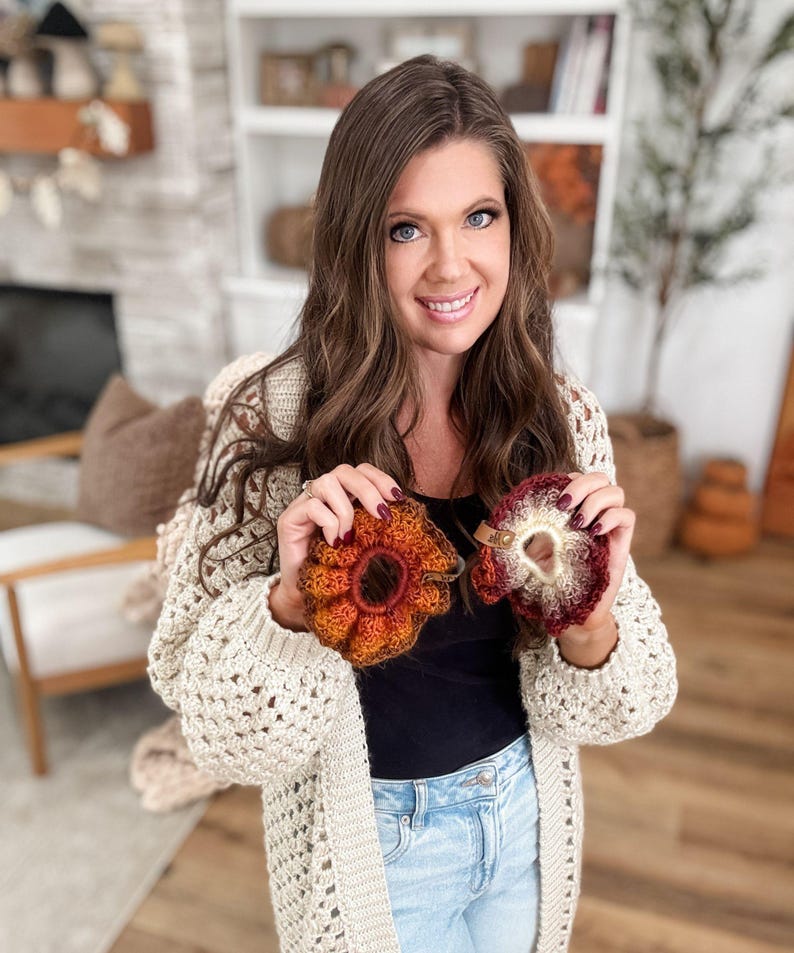 May include: Two handmade crochet hair scrunchies in shades of orange, red, and cream are held up. The scrunchies have a ruffled, flower-like design. The person holding the scrunchies is wearing a cream-colored, open-knit cardigan.