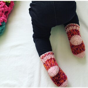 May include: A pair of pink, orange, and red knitted baby socks with a pink circle on the top of each sock.