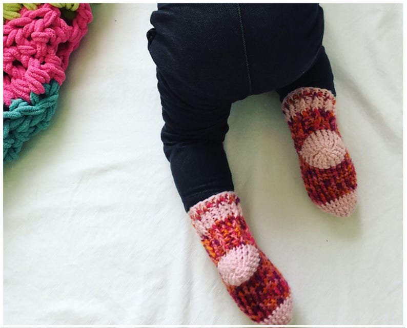 May include: A pair of pink, orange, and red knitted baby socks with a pink circle on the top of each sock.