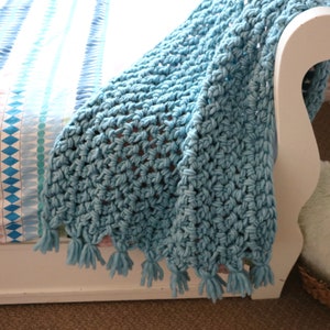Pu&ograve; includere: Una coperta di uncinetto blu chiaro con un bordo a frangia. La coperta &egrave; drappeggiata su una sedia bianca.