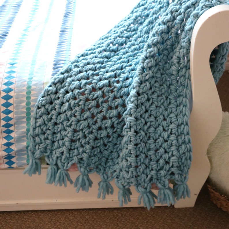Pu&ograve; includere: Una coperta di uncinetto blu chiaro con un bordo a frangia. La coperta &egrave; drappeggiata su una sedia bianca.