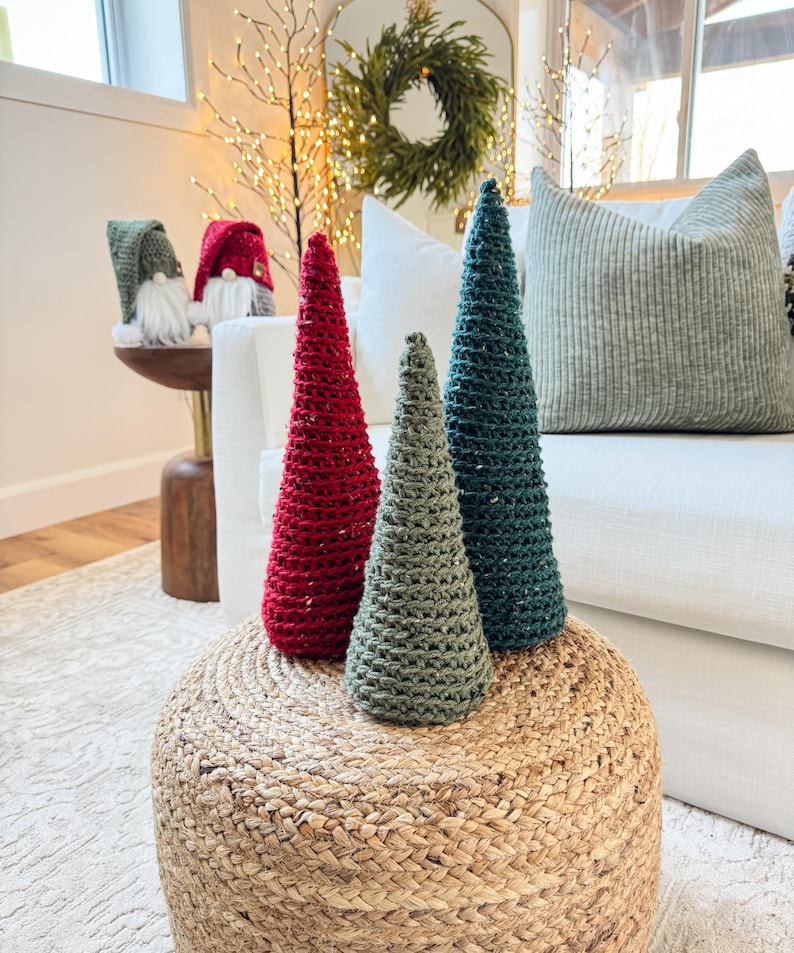 May include: Three crocheted Christmas trees in red, green, and teal colors are displayed on a woven jute ottoman. The trees are arranged in a row from largest to smallest.