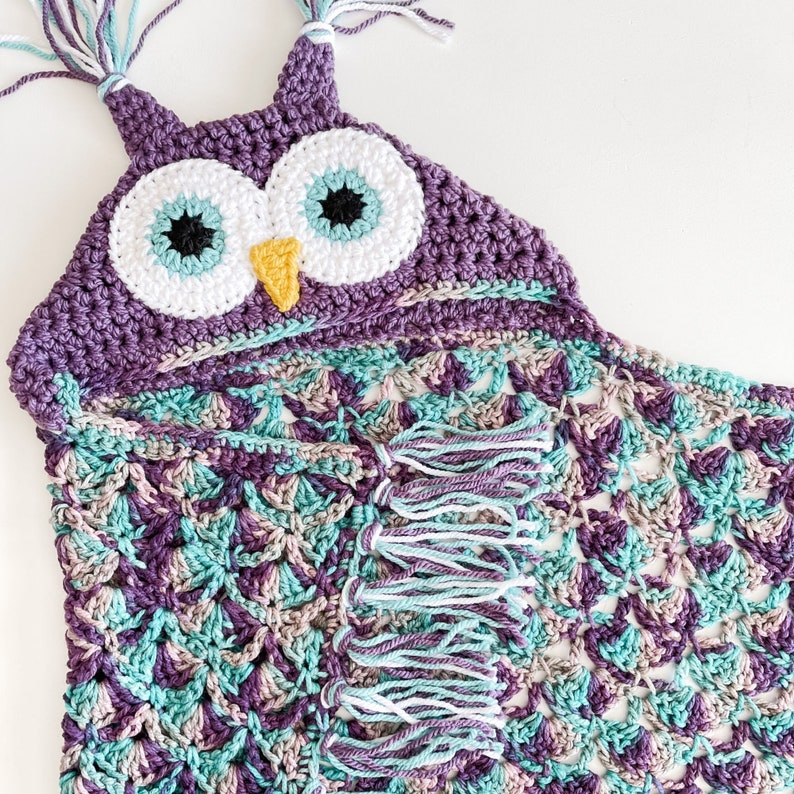 May include: A crocheted owl hooded blanket in shades of purple, teal, and white with a yellow beak and white eyes. The blanket has fringe along the bottom edge.