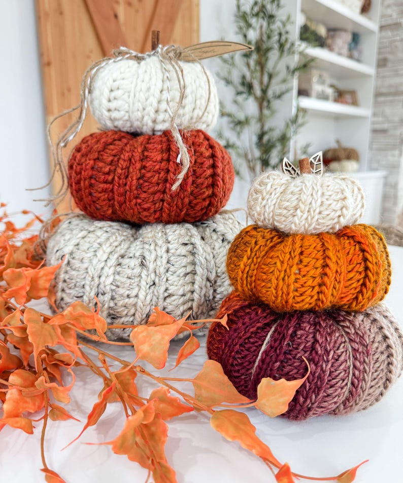K&ouml;nnte beinhalten: Gestapelte, handgestrickte K&uuml;rbisse in Creme, Orange, Rost und Burgunderrot. Jeder K&uuml;rbis hat einen Stiel und ist Teil einer herbstlichen Dekoration.