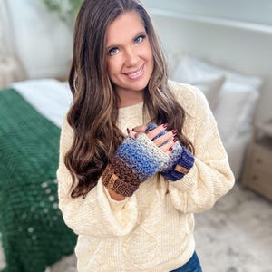 Peut inclure: Mitaines sans doigts crochet&eacute;es dans les tons de marron, bleu et gris. Les gants ont un motif textur&eacute; et de petites &eacute;tiquettes en cuir. La personne porte un pull cr&egrave;me et un jean bleu.
