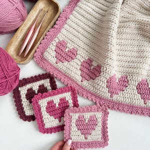 May include: A crocheted blanket with a pink and white color scheme. The blanket has a heart pattern on the border and a heart pattern in the center of each square. The blanket is made with a soft yarn and is perfect for cuddling up on the couch.