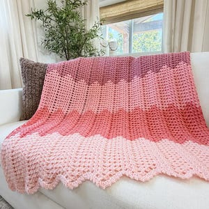 May include: A crochet blanket featuring a chevron pattern with horizontal stripes in shades of light pink, coral, medium pink, and dark mauve. The textile has a scalloped edge and a textured, open-work stitch design throughout the entire piece.