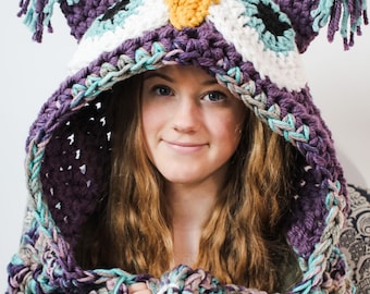 May include: A purple, teal and white crocheted owl hooded scarf with long fringe. The scarf is being worn by a person with long brown hair.