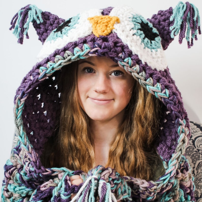 May include: A purple, teal, and white crocheted owl hooded scarf with long fringe. The scarf is worn by a person with long brown hair.