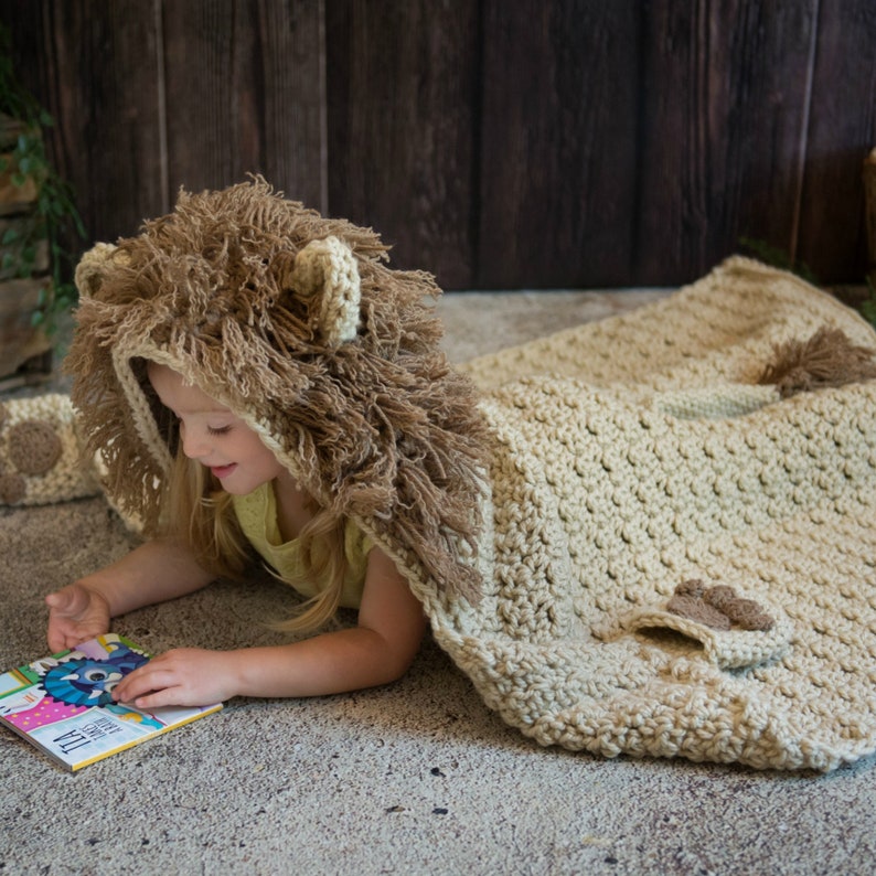 May include: A child wearing a crocheted lion costume with a brown and beige hooded blanket. The blanket has a lion tail and paw prints.