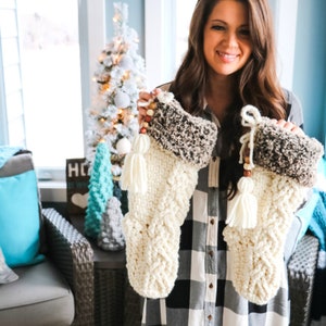 May include: Two white crocheted Christmas stockings with brown faux fur trim and wooden bead accents. The stockings have white tassels and are being held by a person wearing a black and white checked shirt.