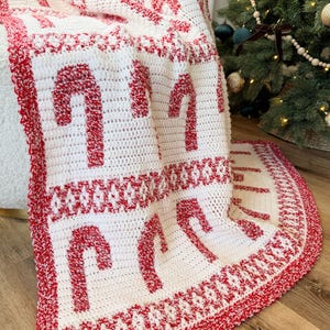 K&ouml;nnte beinhalten: Eine wei&szlig;-rote H&auml;keldecke mit einem Zuckerstangenmuster. Die Decke zeigt gro&szlig;e rote Zuckerstangen auf wei&szlig;em Grund mit einem roten Rand. Die Decke ist &uuml;ber einen Stuhl in der N&auml;he eines Weihnachtsbaums drapiert.