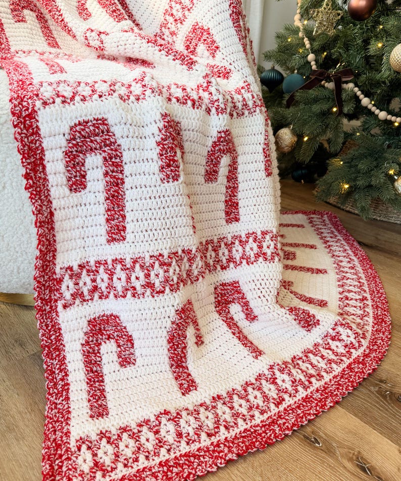 K&ouml;nnte beinhalten: Eine wei&szlig;-rote H&auml;keldecke mit einem Zuckerstangenmuster. Die Decke zeigt gro&szlig;e rote Zuckerstangen auf wei&szlig;em Grund mit einem roten Rand. Die Decke ist &uuml;ber einen Stuhl in der N&auml;he eines Weihnachtsbaums drapiert.