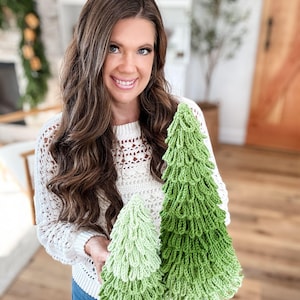 May include: Two handmade crochet Christmas trees in varying shades of green. The trees have a textured, layered design, resembling a fir tree. The larger tree is a vibrant green, while the smaller one is a lighter shade. The trees are displayed against a neutral background.