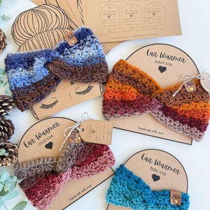 May include: A collection of handmade crochet ear warmers in various sizes and colors. The ear warmers are displayed on cardboard backing with the words "Ear Warmer" and size labels. Colors include blue, brown, red, and orange.