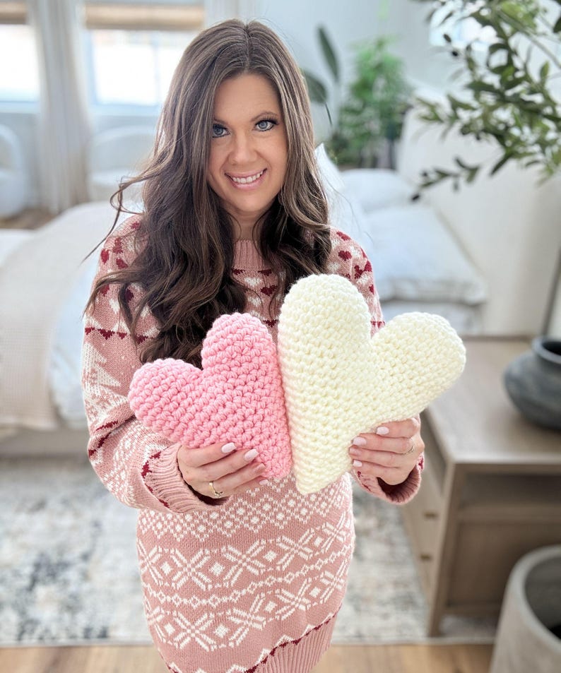 Pu&ograve; includere: Due cuscini a forma di cuore all'uncinetto, uno rosa e uno bianco, tenuti da una persona che indossa un maglione rosa e bianco.