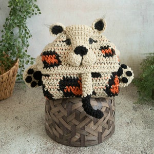 May include: A crocheted tiger hooded blanket with orange, black and white stripes. The blanket is sitting on a brown wicker basket.
