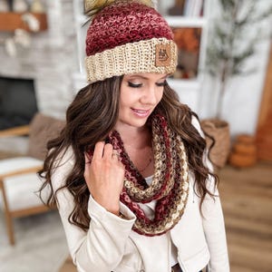 May include: A handmade crochet hat and infinity scarf set in shades of burgundy, cream, and brown. The hat features a faux fur pom-pom and a leather tag. The scarf is a circular design, perfect for cold weather.