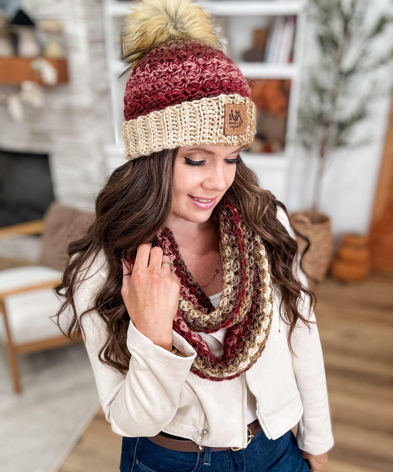 May include: A handmade crochet hat and infinity scarf set in shades of burgundy, cream, and brown. The hat features a faux fur pom-pom and a leather tag. The scarf is a circular design, perfect for cold weather.
